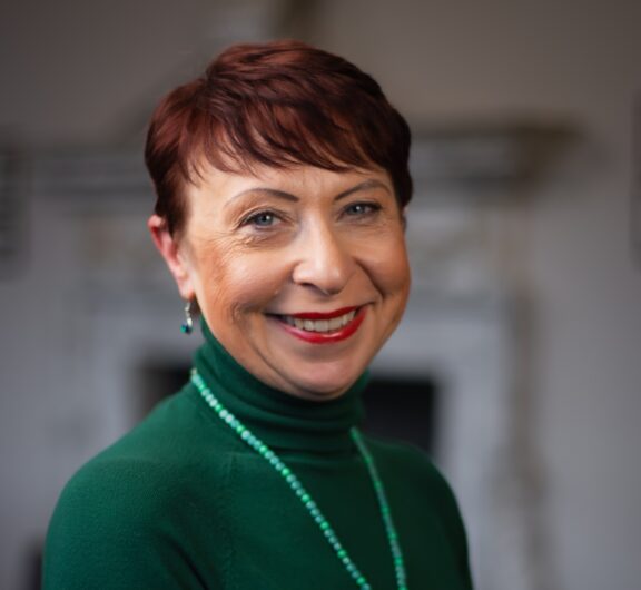 Smiling woman with short red hair, wearing green turtleneck and beads, exuding professionalism.