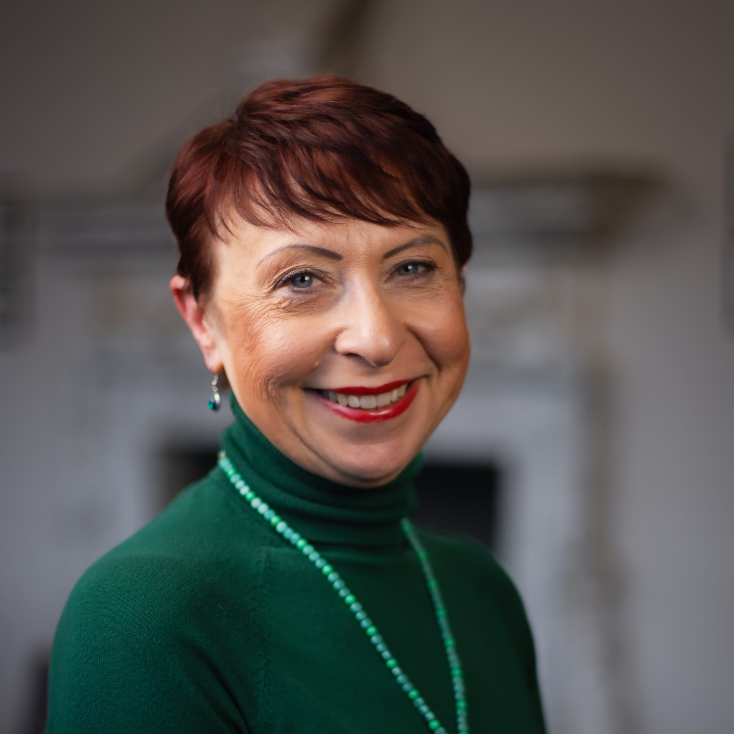 Smiling woman with short red hair, wearing green turtleneck and beads, exuding professionalism.