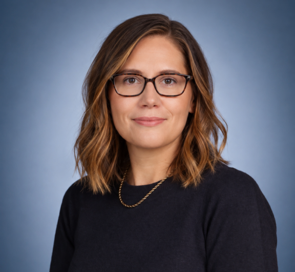 Professional headshot of a woman with glasses and shoulder-length brown hair, wearing a dark blue sweater and a gold necklace against a gradient blue background.