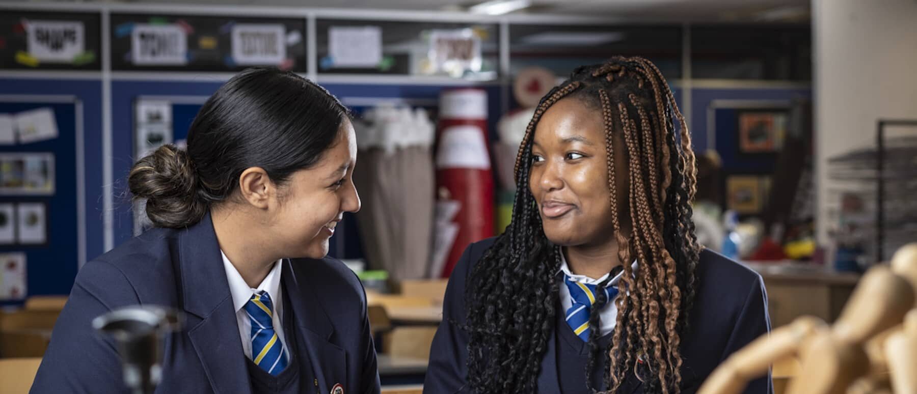 Two students in school uniform discussing in a classroom, promoting student life and educational activities.
