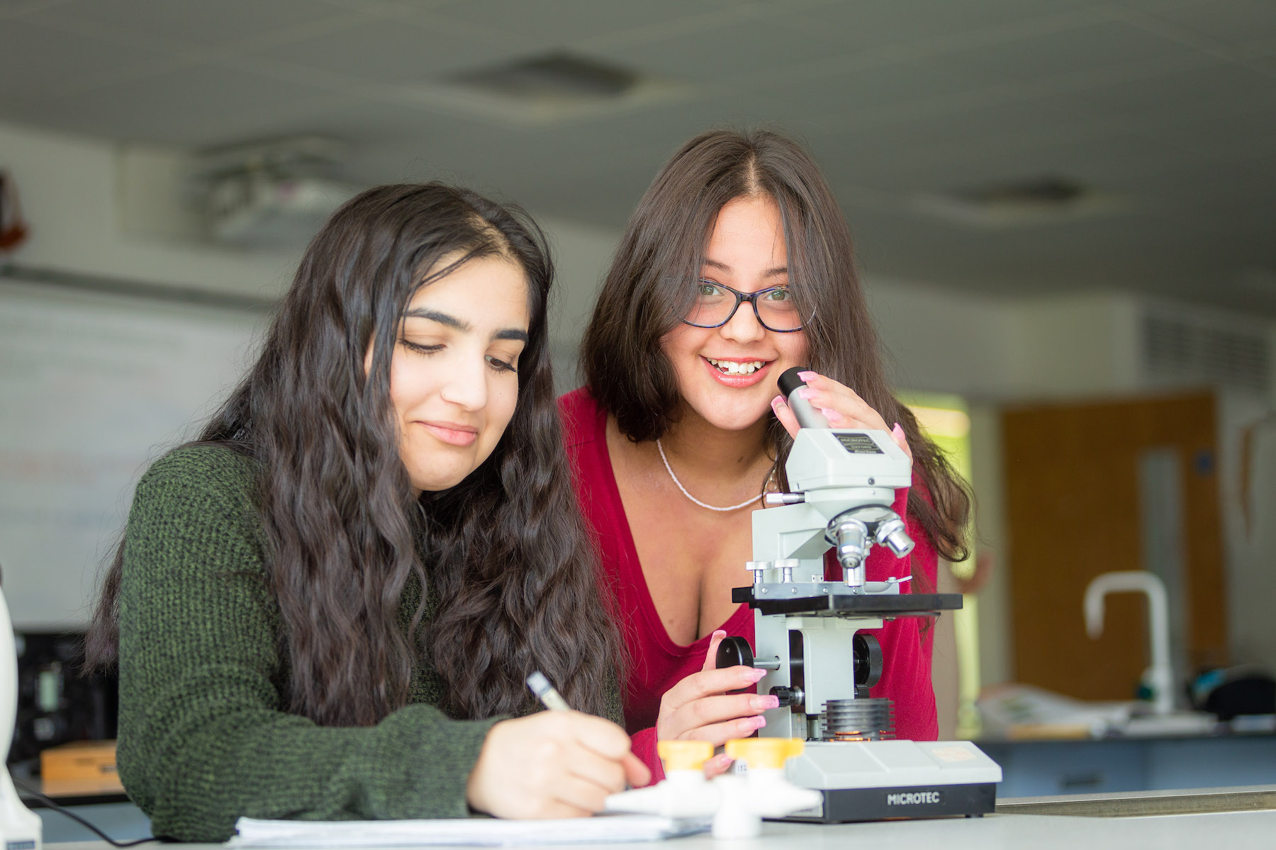 Two female students engaged in a science lesson, one using a microscope while the other takes notes, highlighting educational activities and collaboration.