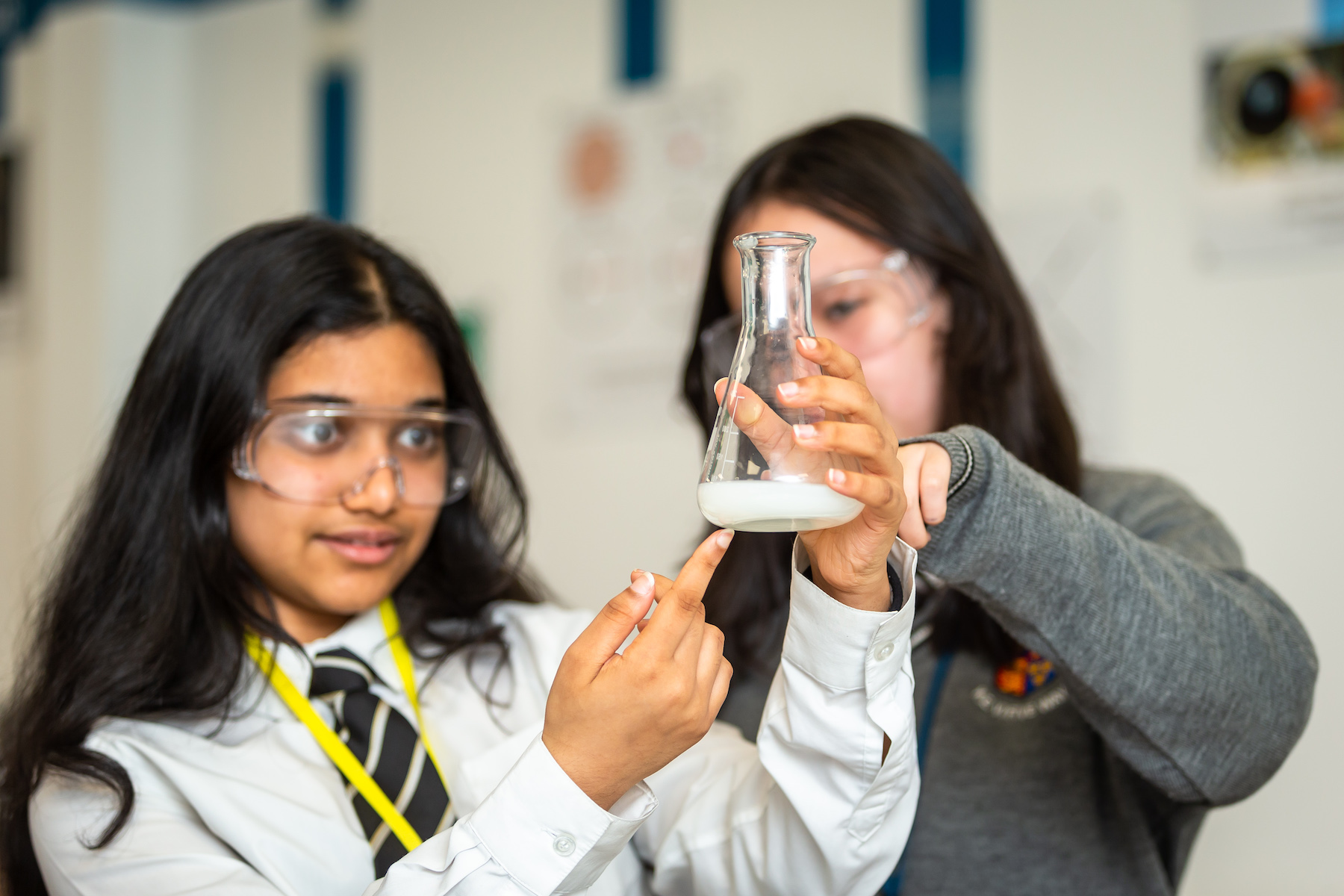 Two students in a science class wearing safety goggles, examining a beaker with a white substance.