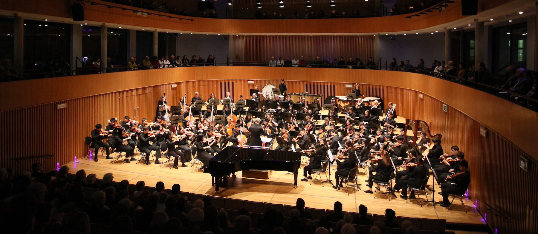 An orchestra performing on a stage in a large auditorium with a conductor leading the musicians.