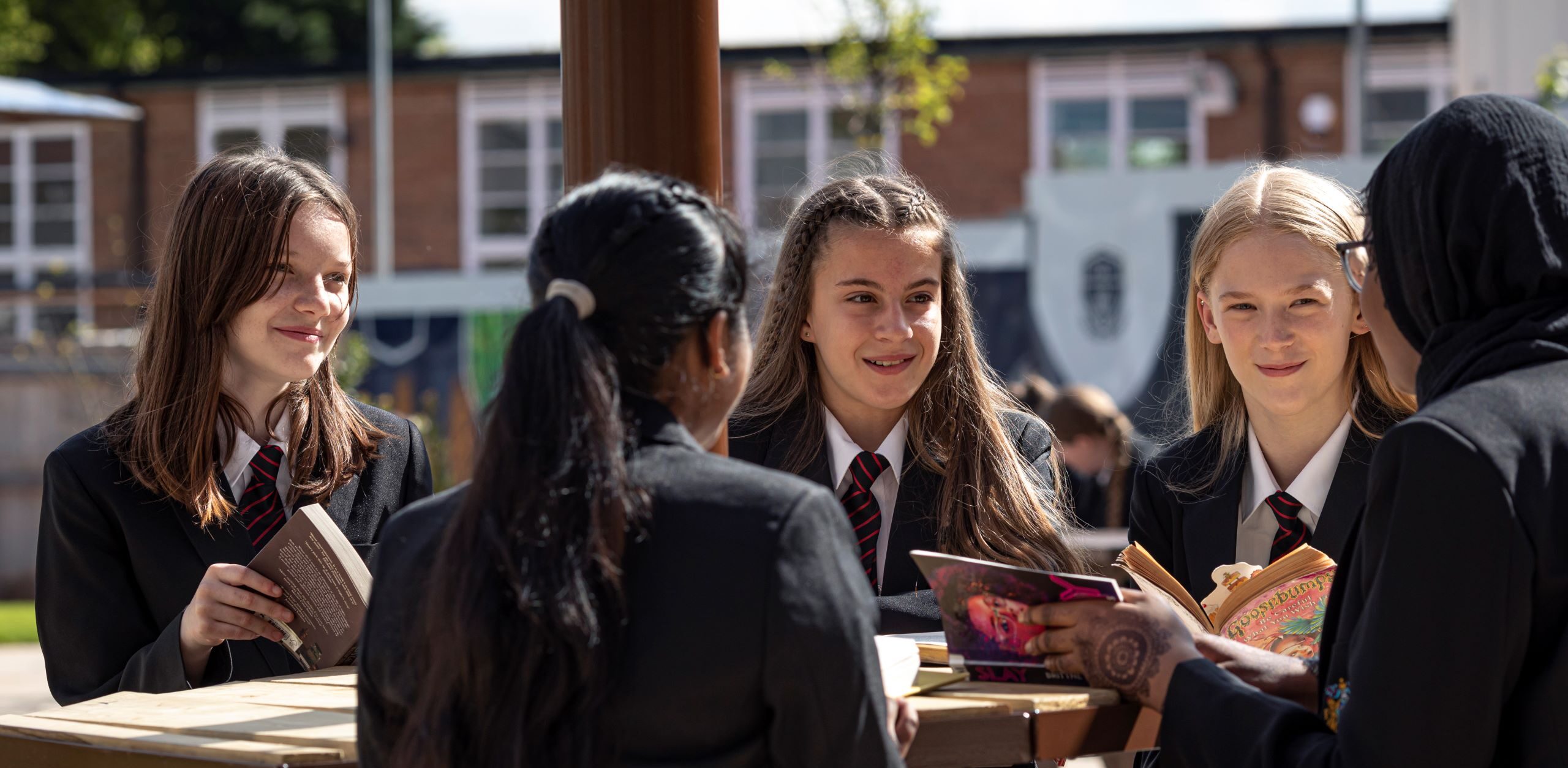 Students in school uniforms sitting at a table outside, engaged in conversation and holding books.