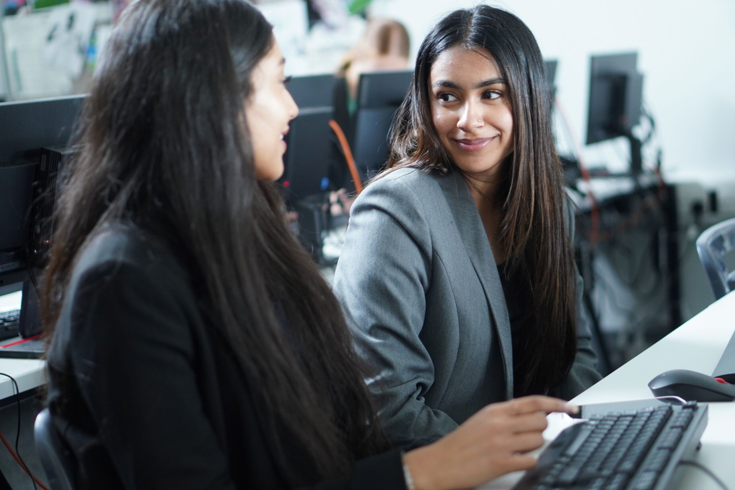 Two young women sitting at a desk, one looking at a computer screen and the other smiling at the camera in a classroom setting.