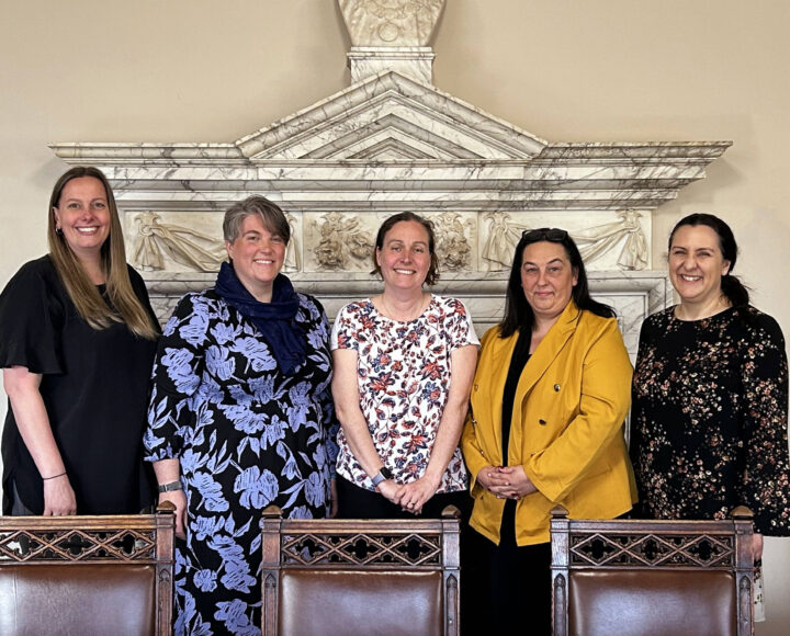 Five women standing together in a formal setting, smiling at the camera, promoting student life and educational activities.