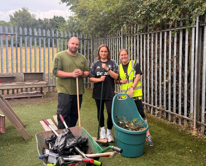 Students in high-vis vests holding cleaning tools next to a wheelbarrow and bin during a school tidy-up activity.