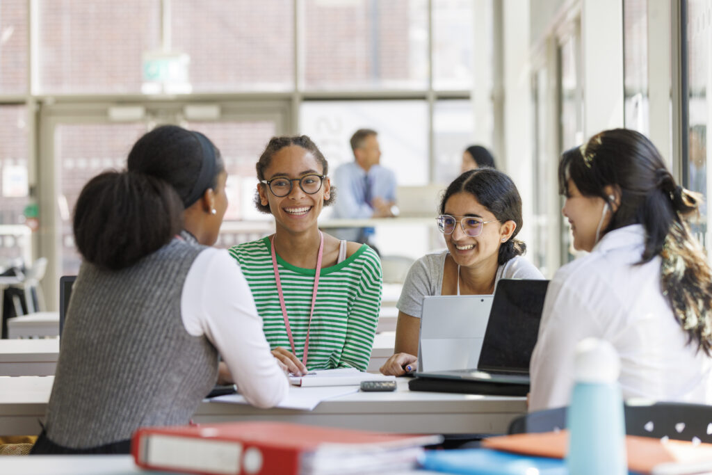 Group of diverse students collaborating at a table in a bright study area. They are smiling and engaged in educational discussion with laptops and textbooks.