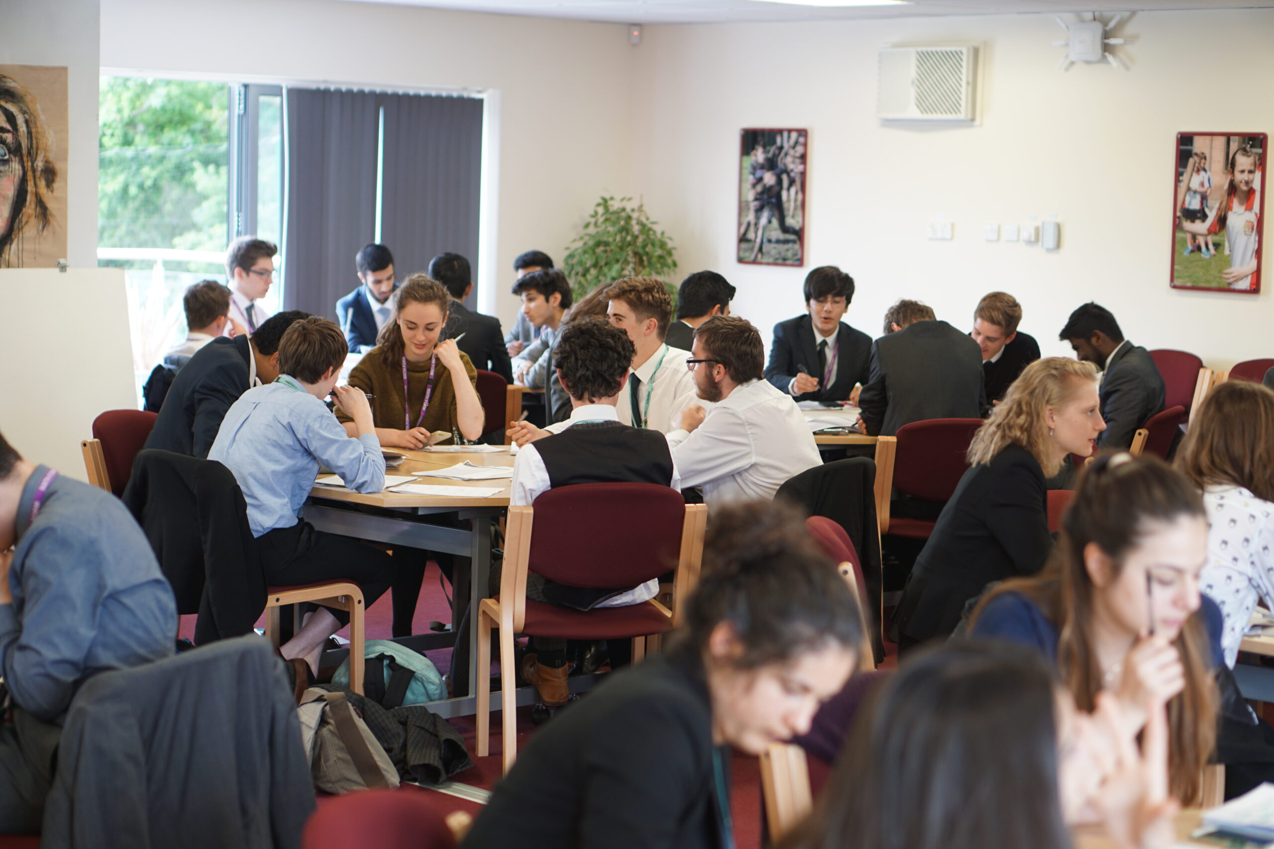 Students in formal attire clustered at group tables within a modern Kevi Foundation seminar room. The collegiate space features natural light, informational posters, and structured seating for collaborative learning.
