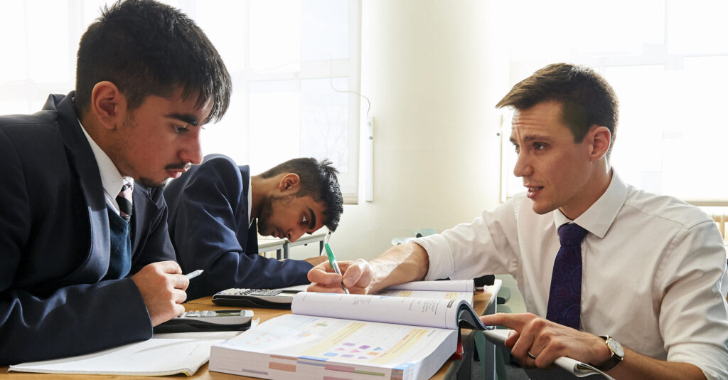 Students and an instructor collaborate over textbooks in a well-lit classroom, reflecting an engaging educational setting. The scene highlights focused study and academic support.